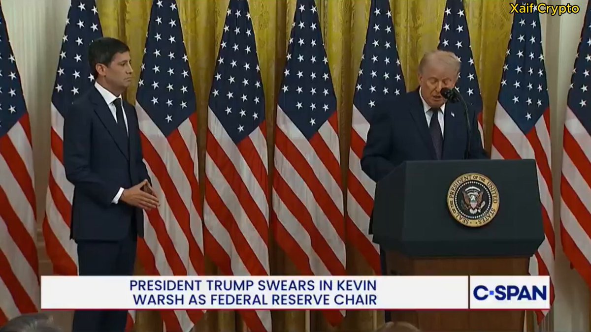 The image shows two men standing at a podium, with one man speaking into a microphone while the other stands behind him. They are dressed in suits and appear to be engaged in a formal event or speech. The background features several American flags, suggesting that this could be an event related to politics or national pride.