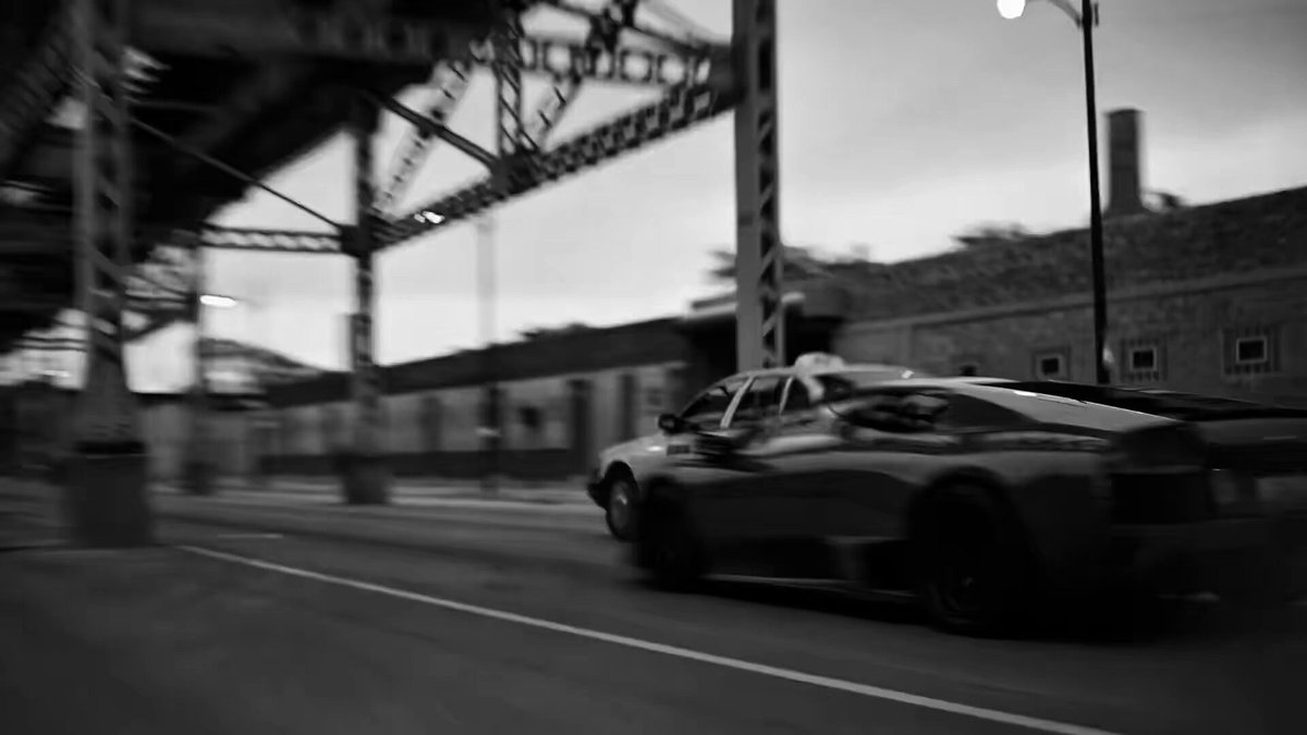 The image shows a car driving down a street under an overpass, with a building visible in the background. The focus of the image seems to be on the car rather than the surrounding environment or other elements present in the scene.