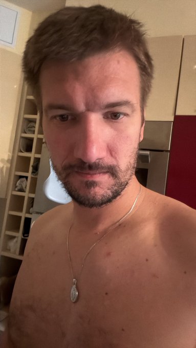A selfie of a shirtless man with short brown hair and a beard, wearing a silver pendant necklace, in what appears to be a home interior.