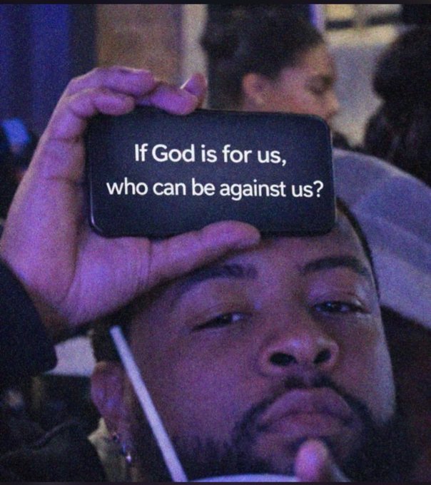 A photo of a man holding up a smartphone displaying the text 'If God is for us, who can be against us?' (Romans 8:31), referencing a popular Kanye West meme.