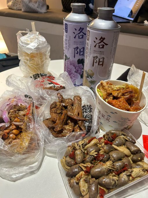 A spread of Luoyang local delicacies rests on a table, including stir-fried sea snails with red chili peppers, braised duck heads/wings, and marinated dishes, accompanied by two cans labeled "洛阳" (Luoyang) featuring floral designs—perfectly matching the caption's celebration of rewarding oneself after a busy day in the city.
