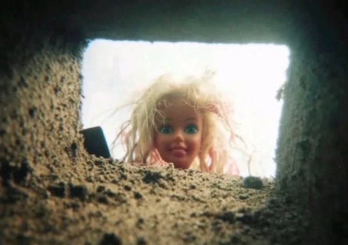 A creepy blonde doll with blue eyes peeking through a hole in a concrete or stone wall, viewed from below looking up.