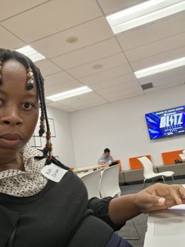 A selfie of a young woman with braided hair in what appears to be a study or library room, with another person visible in the background.