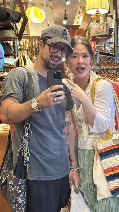 A mirror selfie of a man wearing a grey cap and glasses and a woman with dark hair, both making playful surprised expressions in what appears to be a vintage or thrift store.