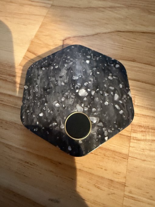 A pentagonal, dark grey speckled object with a small gold-rimmed black circle in the center, resting on a wooden surface in sunlight.