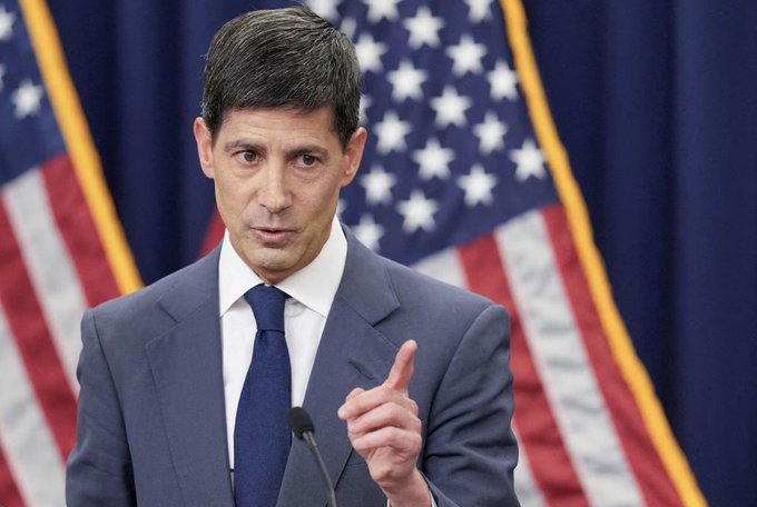 A man in a blue suit gestures while speaking at a podium, with American flags behind him, visually representing the Federal Reserve official referenced in the post about the Fed's recent $27.6 billion Treasury bill purchases signaling renewed balance sheet expansion.