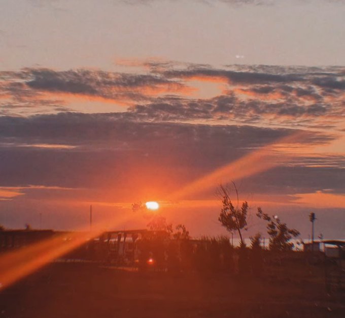 Sunset scene with the sun low on the horizon casting orange light beams through clouds, silhouetting trees and a parked van.