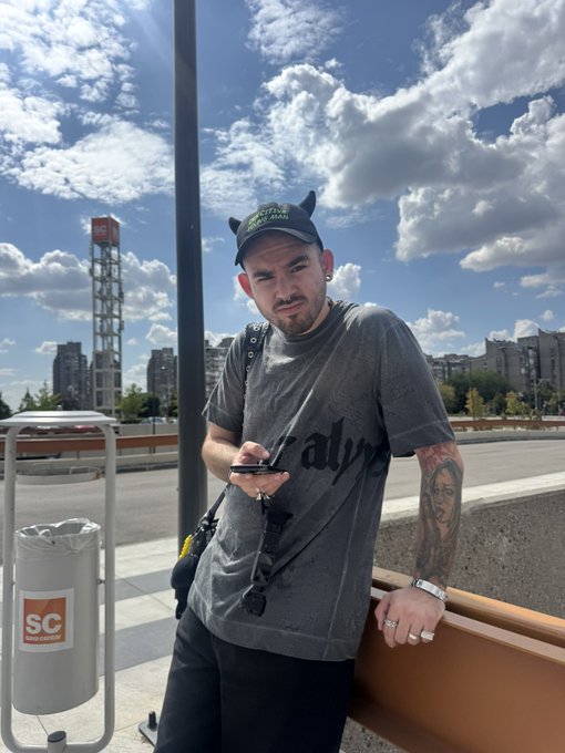 A selfie of a bearded man wearing a black cap with devil horns and a gray t-shirt, standing outdoors with a blue sky and city buildings in the background.
