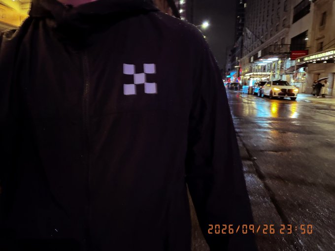 A nighttime street scene shows a person from behind wearing a black jacket with a reflective checkered logo, with a yellow taxi visible on the wet street; the timestamp reads 2026/09/26 23:50.