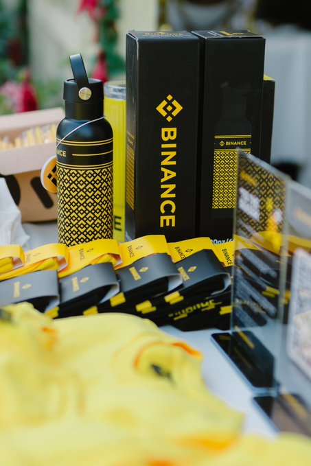 A display of branded Binance merchandise including a black water bottle, boxes with the Binance logo, and yellow lanyards arranged together, perfectly matching the caption's playful nod to a possible excess of Binance-branded items.