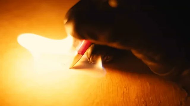 A close-up shot shows a hand holding a pencil, writing on paper illuminated by warm, glowing light against a dark background.
