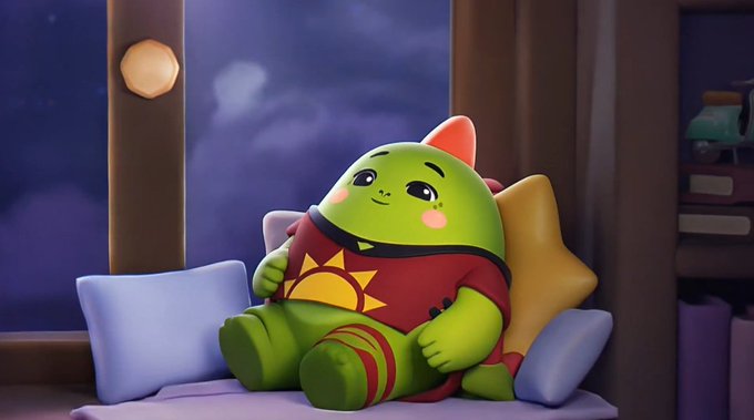 A 3D-rendered cartoon character resembling a green, round creature with a pink bow, wearing a red shirt with a sun design, sitting on a pillow next to a star cushion in a cozy room.