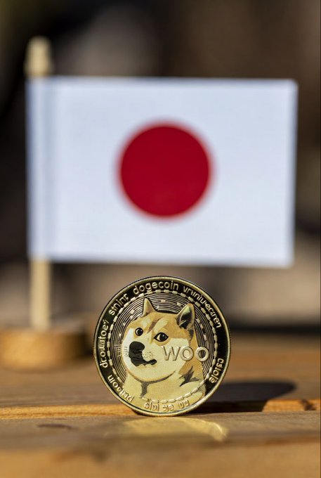 A Dogecoin cryptocurrency coin stands upright on a wooden surface in front of a small Japanese flag, with the coin displaying the iconic Shiba Inu dog and text reading "dogecoin" and "wow" around its edge. The image visually symbolizes Japan's recognition of cryptocurrencies like Dogecoin as legitimate financial assets.