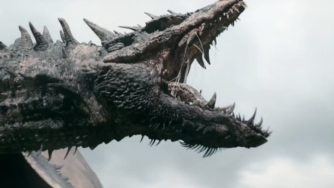 A close-up shot of a dragon's head with sharp teeth and textured scales against a cloudy sky background, appearing to be from a fantasy film or TV series.