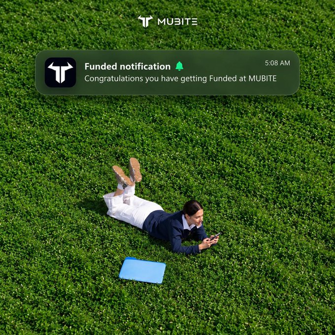 A man in a suit lies on his stomach on grass looking at his phone, with a 'MUBITE' notification reading 'Congratulations you have getting Funded at MUBITE' at 5:08 AM.