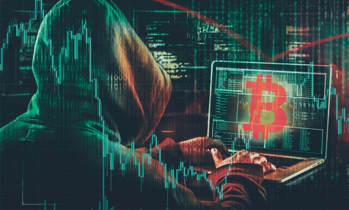 A hooded figure typing on a laptop displaying a red Bitcoin logo, overlaid with green stock/crypto chart candlesticks and binary code.