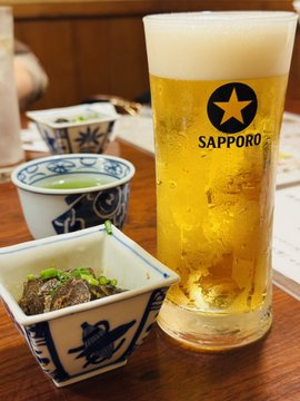 A glass of Sapporo beer sitting on a wooden table alongside small dishes of Japanese food including edamame and what appears to be stewed meat.