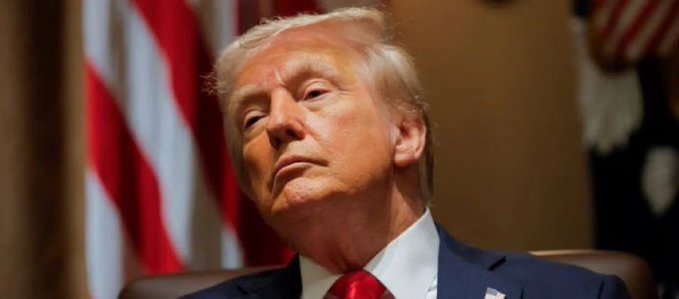 Donald Trump is shown in a suit and red tie, seated in front of an American flag.