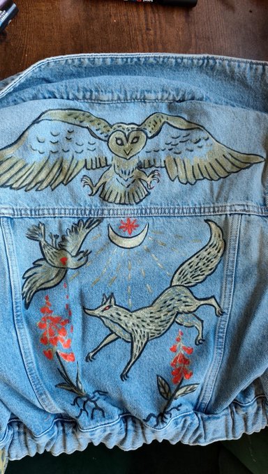 The back of a light blue denim jacket features hand-painted folk art including a large owl, a flying bird, a running fox, a crescent moon, and red flowers.