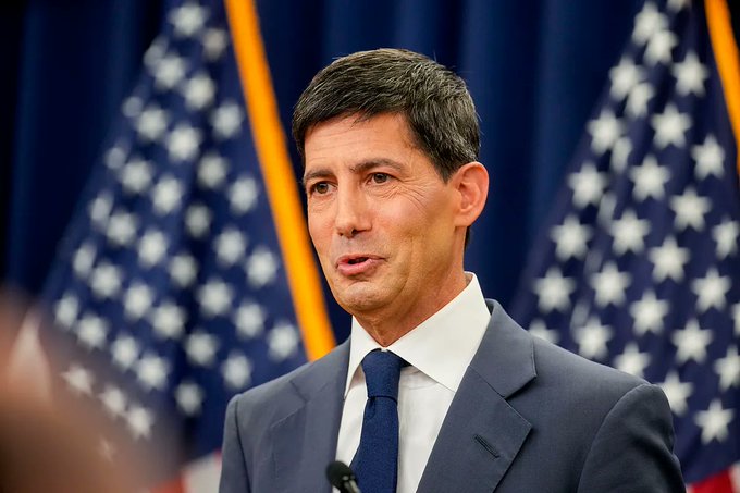 A man in a dark suit and blue tie speaking at a podium with multiple American flags displayed in the background.