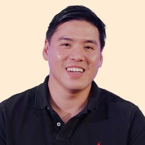 The image features a man wearing a black polo shirt with a red logo, smiling at the camera. The background of the photo is white, which contrasts with the dark tones of the man's outfit. There are no other objects or people visible in the image, and it does not contain any text.
