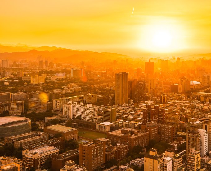 The image shows a cityscape with buildings of various sizes at different heights, including skyscrapers that dominate the skyline. The sun is setting behind these structures, casting an orange glow over the scene. In the foreground, there are two buildings prominently featured - one large building and another smaller one. A person can be seen standing near this larger building, possibly admiring the view or going about their day.