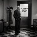 A black and white photograph of a man in a suit standing in front of a door with text that appears to read 'THE BUT... ROOM FOR THE INDIA KITCHEN'.