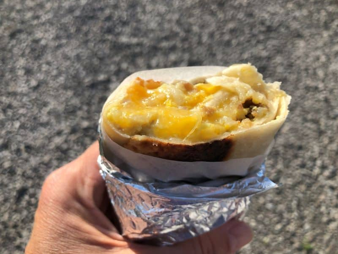 A hand holding a partially eaten burrito wrapped in foil, with melted cheese and egg visible, photographed against an asphalt background.