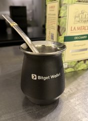 A black metal mate cup with a 'Bitget Wallet' logo and a metal straw sits on a table in front of what appears to be a yerba mate package labeled 'LA MER... DE CAMPO'.