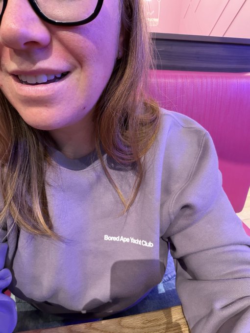 A selfie of a smiling person wearing glasses and a purple 'Bored Ape Yacht Club' sweatshirt, sitting in a booth with pink seating.