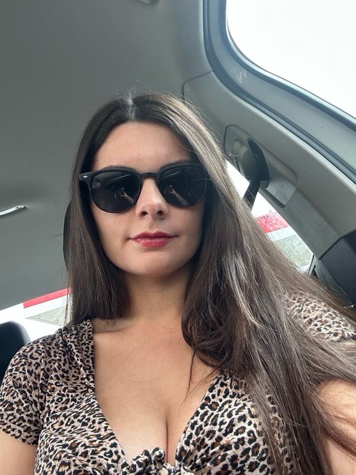 A selfie of a woman with long brown hair wearing black sunglasses and a leopard-print top, taken inside a car.