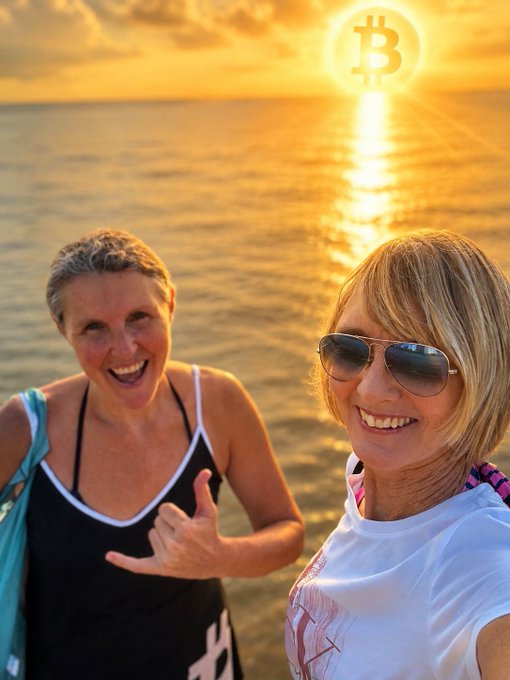 Two women smile for a sunset selfie on a beach, with one giving a shaka hand sign beneath a glowing Bitcoin logo in the sky.