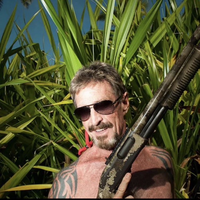A shirtless older man with sunglasses, a mustache, and tattoos holding a rifle, standing in front of tall green plants.