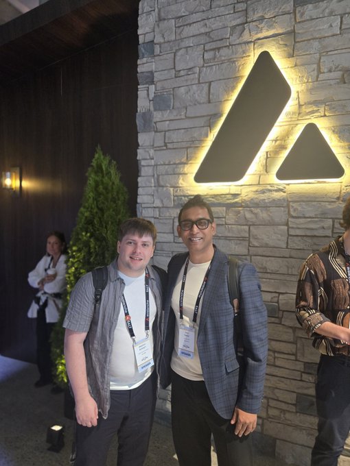 Two men wearing conference lanyards pose together in front of a stone wall with an illuminated logo, at what appears to be a corporate event.