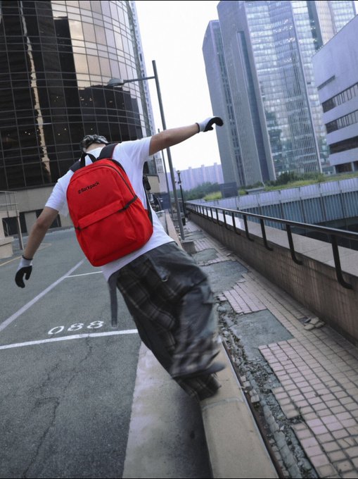 A skateboarder wearing a white t-shirt, plaid pants, and a red 'Supreme' backpack grinds along a curb in an urban setting with modern glass buildings in the background.