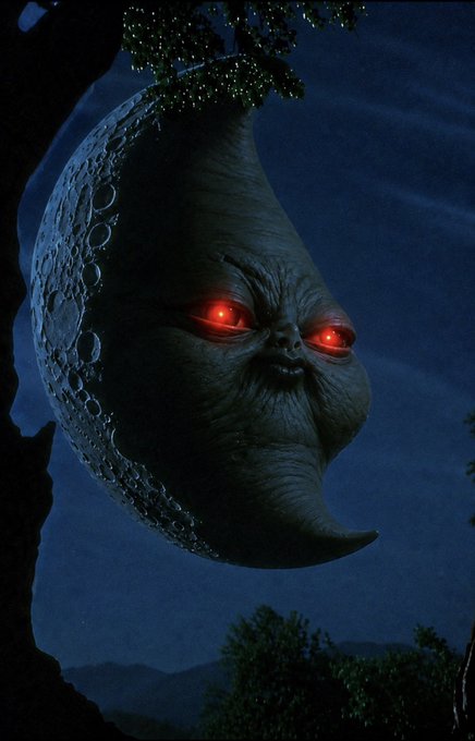 A menacing crescent moon with a face and glowing red eyes hangs in a dark night sky, appearing to be from a fantasy or horror film scene with trees framing the composition.