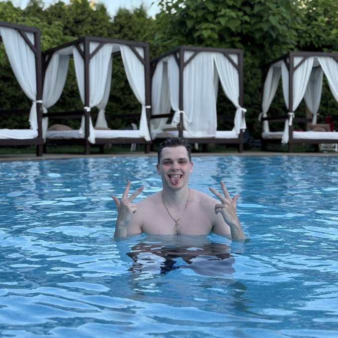 A young man smiles and makes peace signs while partially submerged in a blue swimming pool, with white-curtained cabanas lined up behind him.