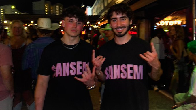 Two smiling men pose at a crowded nighttime event, both wearing matching black t-shirts with "$ANSEM" printed in pink lettering, representing the ticker. A "COYOTE" sign glows in the background as they throw up hand gestures to show their support.
