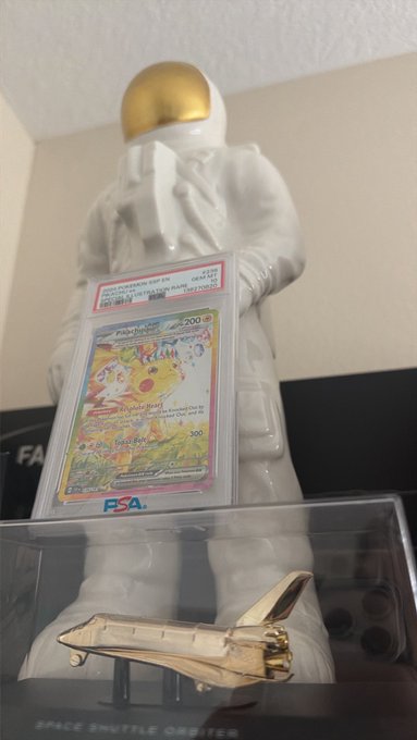 A PSA-graded Pokémon card displayed on a clear stand in front of a white astronaut figurine, with a gold Space Shuttle Orbiter model visible below.