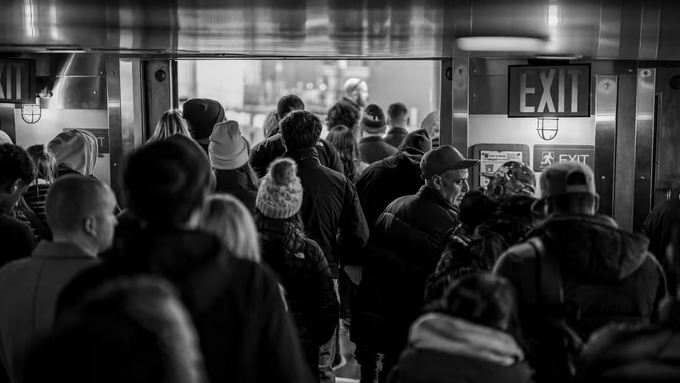 A black-and-white photograph of a crowded indoor space, likely a subway station, with many people gathered near an illuminated "EXIT" sign.
