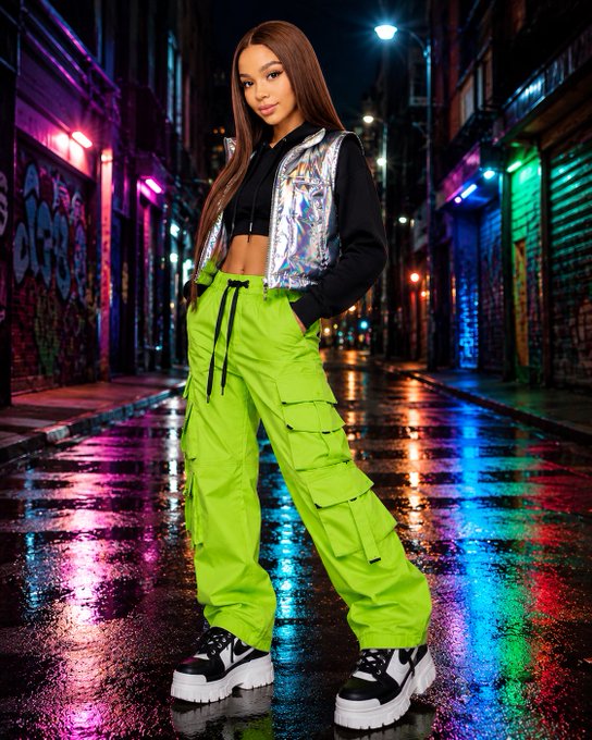 The image features a woman wearing neon green pants, standing on a wet city street at night. She has her arms crossed over her chest and is posing for the camera with a serious expression. The background of the photo includes colorful graffiti-covered buildings that add an urban vibe to the scene. There are also neon lights in the background, which further enhance the nighttime atmosphere.