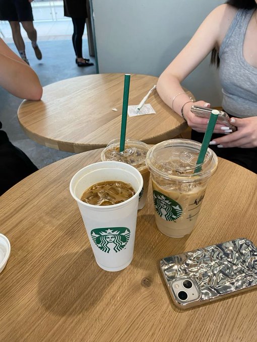 A photo of three Starbucks iced coffee drinks on a wooden table, with one empty cup and a phone with a crumpled foil case, while a person in gray sits nearby using their phone.