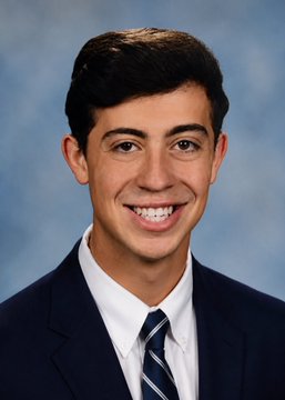 A young man with dark hair smiles at the camera in a formal headshot, wearing a dark suit, white collared shirt, and striped navy tie against a blue backdrop. The image appears to be a professional portrait, likely unrelated to the post mentioning "two floor cryptopunks" transactions.