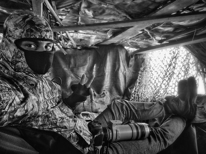 A black and white photograph of a photographer wearing a camouflage balaclava and gear, sitting inside a camouflaged blind while holding a camera with a large Canon telephoto lens, making a peace sign.