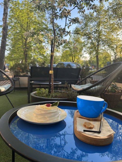 A blue coffee cup and a small white dessert with a raspberry sit on a wooden tray on a blue glass table at an outdoor café, with trees and seating visible in the background.