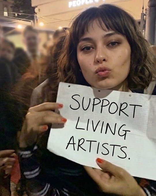 The image features a woman holding up a sign that reads "support living artists." The background of the photo shows people walking by, indicating an outdoor setting or event where this message might be relevant. The caption of the image also mentions an auction starting at 0.50 ETH and is related to the post in some way.