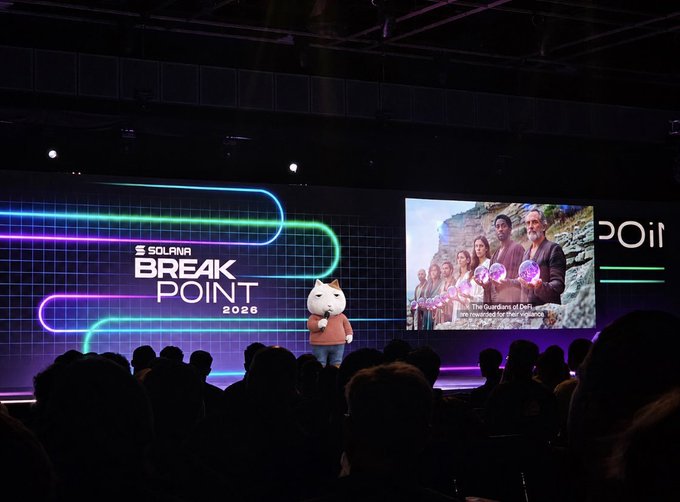 A photograph from the Solana Breakpoint 2026 conference showing a speaker wearing a white cat-head mask on stage in front of a large screen.