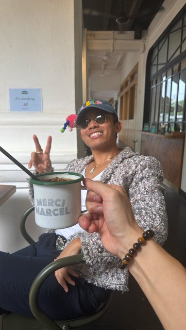 A smiling person wearing sunglasses and a colorful cap holds up a peace sign while seated at what looks like a café, with another person's hand in the foreground holding a cup labeled 'MERCI MARCEL'.