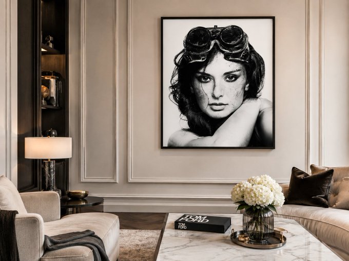 An elegant living room interior with a large framed black-and-white portrait of a woman wearing aviator goggles on the wall, alongside a sofa, coffee table with flowers, and a Tom Ford book on the floor.
