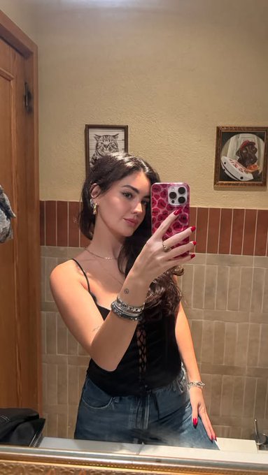 The image shows a woman taking a selfie with her cell phone, capturing herself in front of a mirror. The photo includes an overlay of a Bitcoin symbol, which might be related to the ongoing discussion about Bitcoin's stability or support. The presence of this symbol suggests that the woman is interested in cryptocurrency and its market trends.